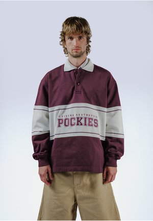 Jeune homme aux cheveux châtain clair portant un polo à manches longues violet et blanc avec le texte "POCKIES", debout devant un fond clair uni.
