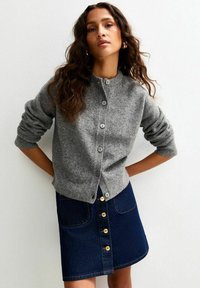 Grijze wollen cardigan met ronde hals en knoopsluiting, gecombineerd met een donkerblauwe denim A-lijn rok met gouden knopen en zakken.
