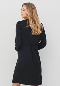 Černé šaty s dlouhými rukávy a elastickým pasem, vyrobené z hladké látky a s uvolněným střihem. Viditelný detail bočních kapes.