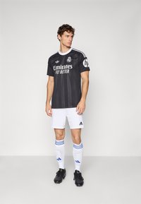 Man wearing black and white Real Madrid soccer kit, standing with legs slightly apart, looking to his left against a plain background.