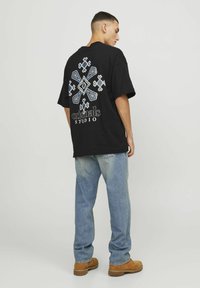 Maglietta nera oversize con un motivo geometrico bianco e blu sul retro, con il testo nero "originals STUDIO" sotto il design. Indossata con jeans chiari.