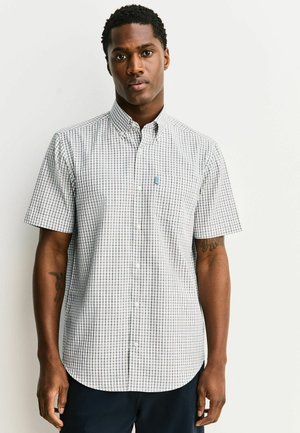 Young man with short curly hair wearing a white and gray checked short-sleeve button-up shirt standing against a plain background.