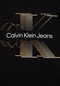 Černé tričko má centrální logo se stylizovaným 'CK' ve zlaté a bílé barvě, překrývající pruhy a text "Calvin Klein Jeans" pod tím.