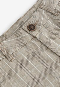 Pantalons à carreaux beiges en tissu texturé, avec des détails de bouton marron et des passants de ceinture, présentant un motif en grille avec des rayures contrastées.