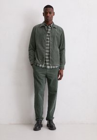 Zielona sztruksowa kurtka i pasujące spodnie, oba z pionowym prążkowaniem, noszone na koszuli w kratę z zielonym i białym wzorem. Czarne buty.