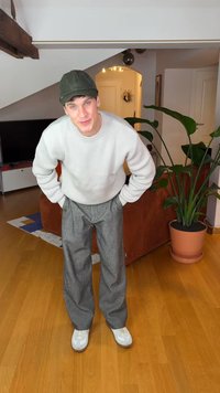 Helehall halli kampsun, lahtised hallid ruudulised püksid ja valged tossud. Roheline nokamüts lõpetab komplekti. Siseruumides taim taustal.