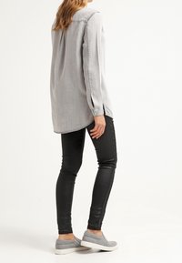 Camicia grigia chiaro a maniche lunghe con polsini a bottone, abbinata a leggings neri e scarpe slip-on grigie; tessuto liscio e vestibilità ampia.