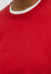 Personne portant un pull à col rond rouge sur une chemise à col rond blanche, recadré pour montrer le col et le haut de la poitrine.