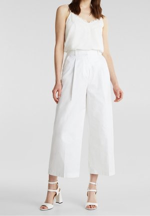 Femme portant un caraco blanc orné de dentelle, un pantalon blanc à jambes larges taille haute, et des sandales blanches à talons blocs avec brides, debout sur un fond gris.