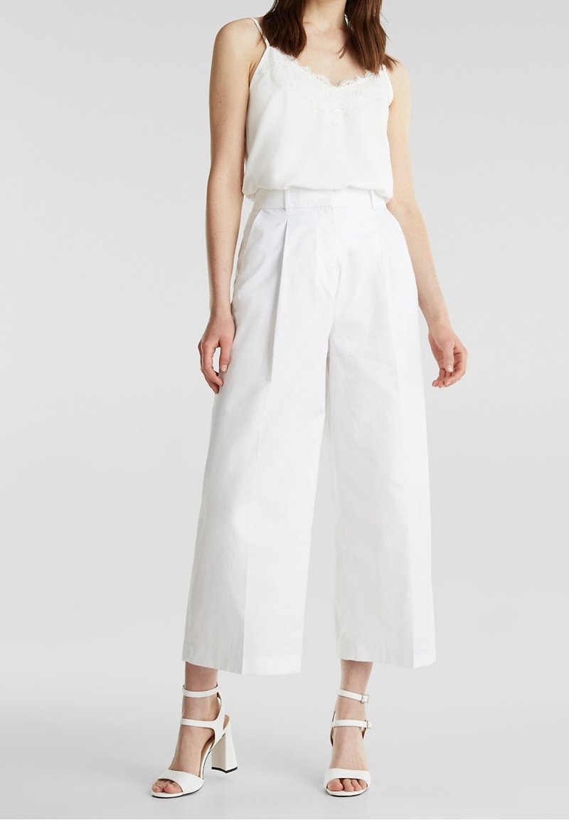 Femme portant un caraco blanc orné de dentelle, un pantalon blanc à jambes larges taille haute, et des sandales blanches à talons blocs avec brides, debout sur un fond gris.