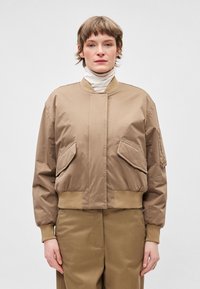 Tan bež jakna bomber z gladkim zaključkom, elastičnimi manšetami, dvema velikima žepoma spredaj in rebrastim pasom. Nošena čez majico z visokim ovratnikom.