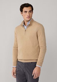 Uomo che indossa un maglione beige con zip sopra una camicia a righe blu e pantaloni grigio scuro, in piedi contro uno sfondo grigio chiaro uniforme.