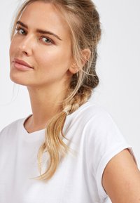 T-shirt en coton blanc avec des manches retroussées, doté d'une coupe décontractée et d'une texture douce, porté par une personne aux cheveux tressés.