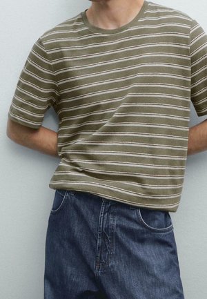 Homme portant une chemise à manches courtes rayée olive et blanche avec un jean en denim bleu, mains derrière le dos.