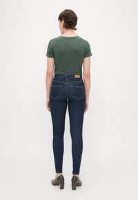 Jeans skinny blu scuro a vita alta, con tasche posteriori e un'etichetta beige. Abbinati a una maglietta verde a maniche corte e tacchi marroni.