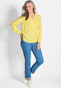 Gelbe Langarmbluse mit Henley-Ausschnitt und fünf Knöpfen, kombiniert mit blauen geraden Jeans und weißen Sneakers.