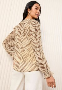 Blusa beige con motivo ondulato marrone, maniche lunghe a palloncino e polsini elasticizzati. Tessuto leggero con un design fluido.