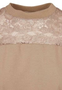 Beige Oberteil mit Rundhalsausschnitt, das einen Spitzenbereich über den Schultern aufweist. Glatter Stoff unten mit einem strukturierten Spitzenoberteil.