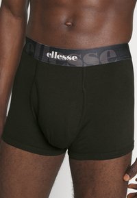 Schwarze Baumwollboxershorts mit niedrigem Bund und elastischem Taillenbund, der das "ellesse"-Logo in Weiß und ein halbtransparentes Muster aufweist.