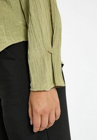 Camicia a maniche lunghe verde chiaro, sgualcita, con chiusura a bottone ai polsi, abbinata a pantaloni neri. Tessuto morbido e vestibilità comoda.