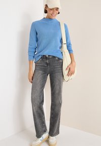 Hellblauer, strukturierter Pullover, graue Jeans, cremefarbige Umhängetasche und weiße Sneaker mit blauen Akzenten. Leger Outfit vor einem neutralen Hintergrund.