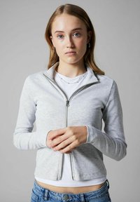 Giacca grigia con zip e texture a coste, dotata di colletto alto e maniche lunghe, indossata sopra una camicia bianca. Parte inferiore in denim casual.