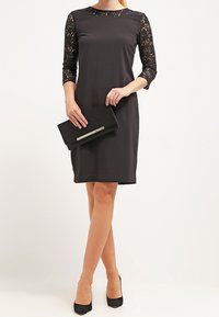 Černé šaty s třičtvrtinovými krajkovými rukávy, kulatým výstřihem, rovnou siluetou a délkou po kolena, doplněné černou kabelkou.