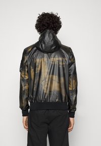 Svart huva jacka med en glänsande, texturerad yta och bruna abstrakta mönster. Har en ribbad nederkant och ärmar, samt en sidoficka.