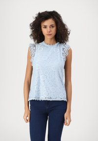 Ανοιχτό μπλε τοπ από δαντέλα με κυματοειδείς μανσέτες και στρίφωμα, διακοσμημένο με λουλουδάτα μοτίβα και πολύπλοκες λεπτομέρειες. Συνδυάζεται με σκούρε�ς τζην παντελόνες.