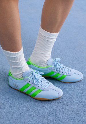 Pieds portant des baskets bleu clair avec des bandes vert vif et des chaussettes blanches côtelées, debout sur un tapis bleu.