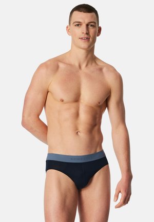 Joven con cabello corto que lleva puestos calzoncillos negros con una cintura gris, de pie contra un fondo claro y liso.