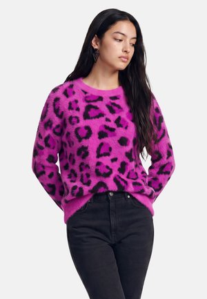 Vrouw met lang donker haar die een felroze, pluizige trui met zwarte luipaardvlekken en zwarte broek draagt, staand met handen in de zakken.