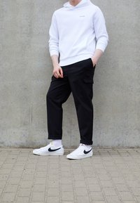 Witte hoodie met Calvin Klein-logo, zwarte cargobroek met zijzakken, en witte sneakers met zwarte swoosh op een gestructureerde grijze achtergrond.