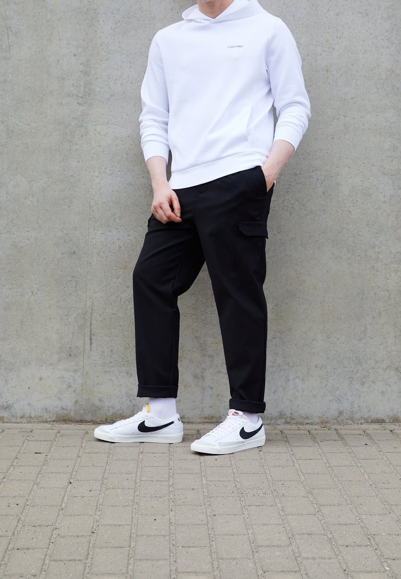 Weißer Kapuzenpullover mit Calvin Klein-Logo, schwarze Cargo-Hosen mit seitlichen Taschen und weiße Sneaker mit schwarzem Swoosh auf strukturiertem grauem Hintergrund.