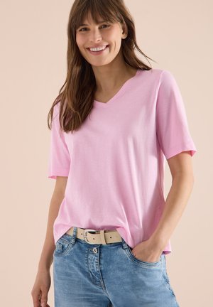 Femme souriante aux longs cheveux bruns portant un t-shirt rose clair à col en V, un jean bleu et une ceinture beige, une main dans la poche.