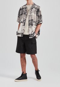Mężczyzna w czarnych szortach cargo, czarnych sneakersach za kostkę, kremowej koszulce i luźnej, krótkosleeve'owej koszuli zapinanej na guziki z abstrakcyjnym czarno-białym wzorem.