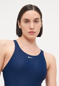 Mørkeblå svømmedrakt har en rund hals og tykke stropper, med en liten hvit Nike-logo på brystet. Materialet har en jevn tekstur.