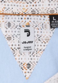 Geweven stof met een geometrisch patroon in oranje, zwart en roomkleur; bevat een lichtblauw gedeelte en een label met de tekst "J&JOY, Ontworpen in België."