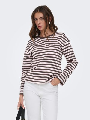 Kobieta ubrana w brązowo-białą pasiastą bluzkę z długim rękawem i białe spodnie, trzymająca w lewej ręce czarną torbę na pasku.