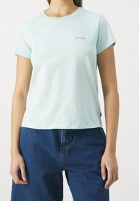 Minit vihreä puuvillapaita lyhyillä hihoilla, jossa on vaaleanpunainen brodeerattu Levi's-logo vasemmassa rintakehässä. Rentomallinen ja sileä rakenne.