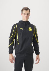 Chaqueta deportiva negra con rayas amarillas, con capucha, cremallera frontal y logo del BVB. Fabricada en tejido ligero, con puños texturizados.