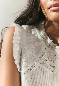 White blouse featuring pleated fabric, floral embroidery, and lace trim at the collar and shoulders. Button closure visible at the front.