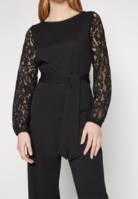 Svart jumpsuit med långa, genomskinliga spetsärmar och en figurnära midja. Har ett matchande bälte och en enkel rund halsringning.