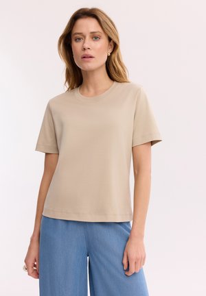 Kobieta o jasnobrązowych włosach, ubrana w beżową koszulę z krótkim rękawem i niebieskie spodnie, stoi na jednolitym jasnym tle.