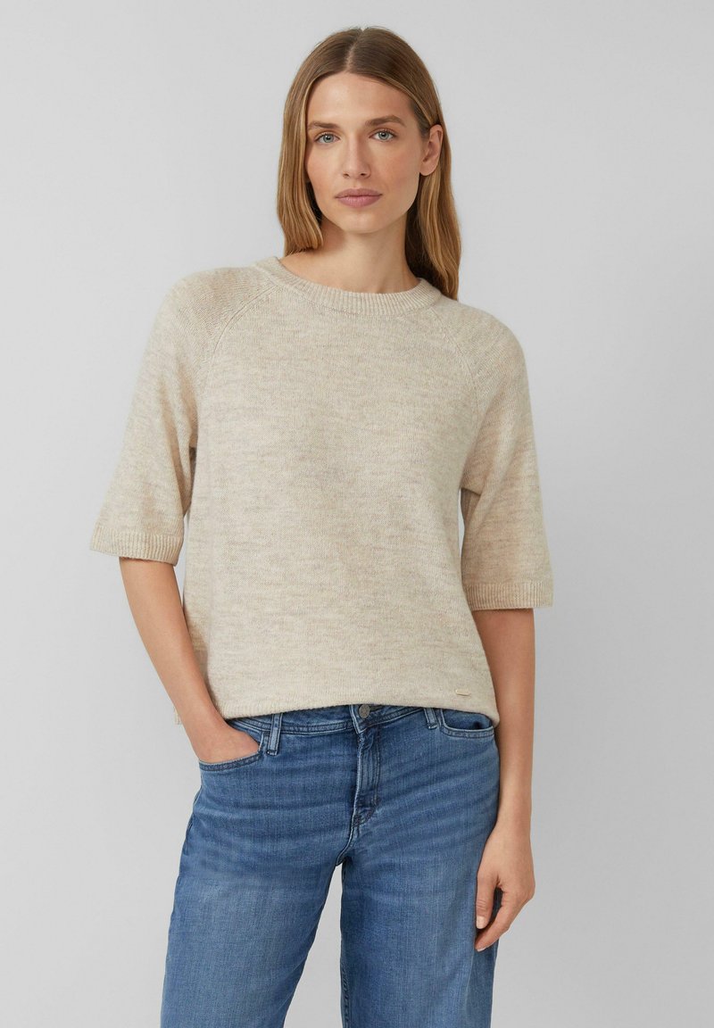 Suéter de punto beige con mangas cortas y cuello redondo, combinado con jeans azules. Textura suave, diseño casual, con detalles de costura visibles.