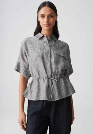 Femme portant une chemise en lin gris à manches courtes avec un nœud à la taille à l'avant et un pantalon noir, debout devant un fond clair uni.