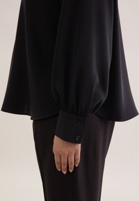 Blusa nera con maniche lunghe e puffate, dotata di un polsino con due bottoni decorativi. Il tessuto appare liscio e leggero.