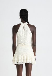 Una donna con i capelli intrecciati che indossa un abito senza maniche in pizzo bianco testurizzato con un orlo arricciato, vista di spalle su uno sfondo semplice.