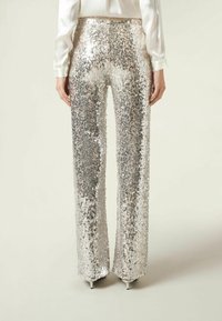 Kobieta ubrana w szerokie spodnie z wysokim stanem, pokryte srebrnymi cekinami, oraz białą bluzkę, stojąca na tle jasnego, jednolitego tła.