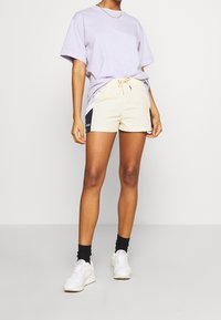 Fialové tričko s krátkým rukávem, béžové šortky s černými akcenty, stahovací pas, doplněné bílými teniskami a černými kotníkovými ponožkami.